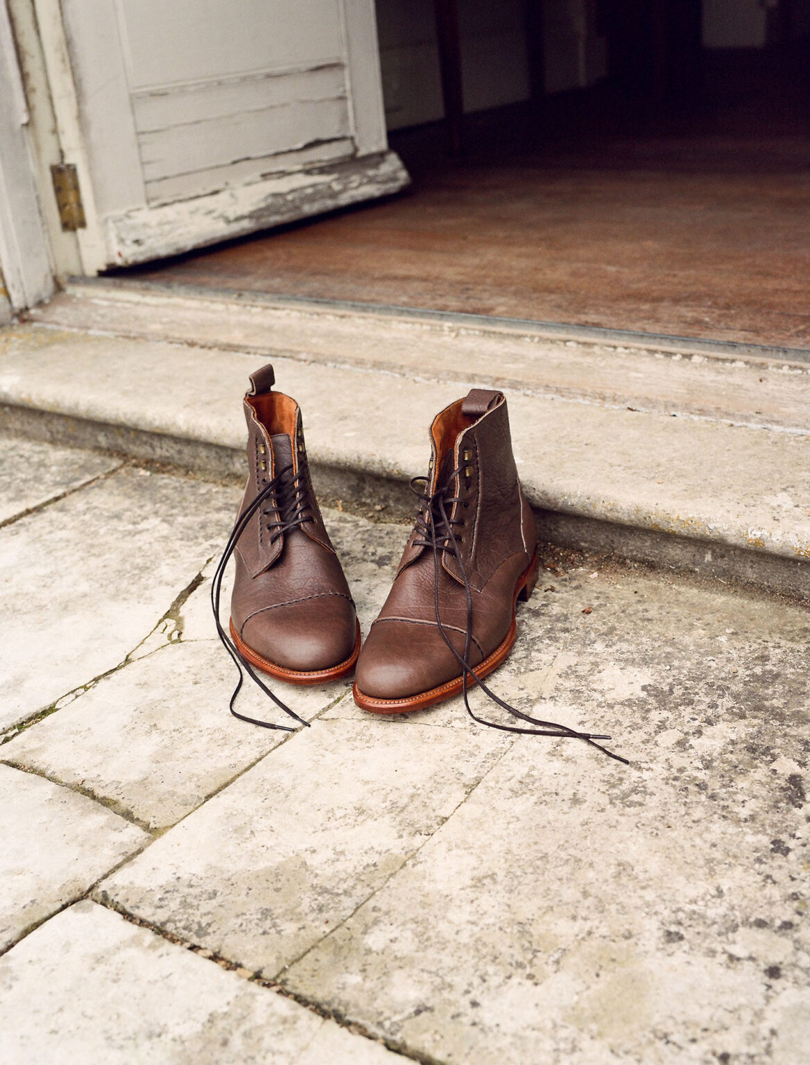 A pair of E.Woodford Barrack 'Stanley' WWII Boots in Grey Horween Veg Tanned Bison standing on a patio outside an open door