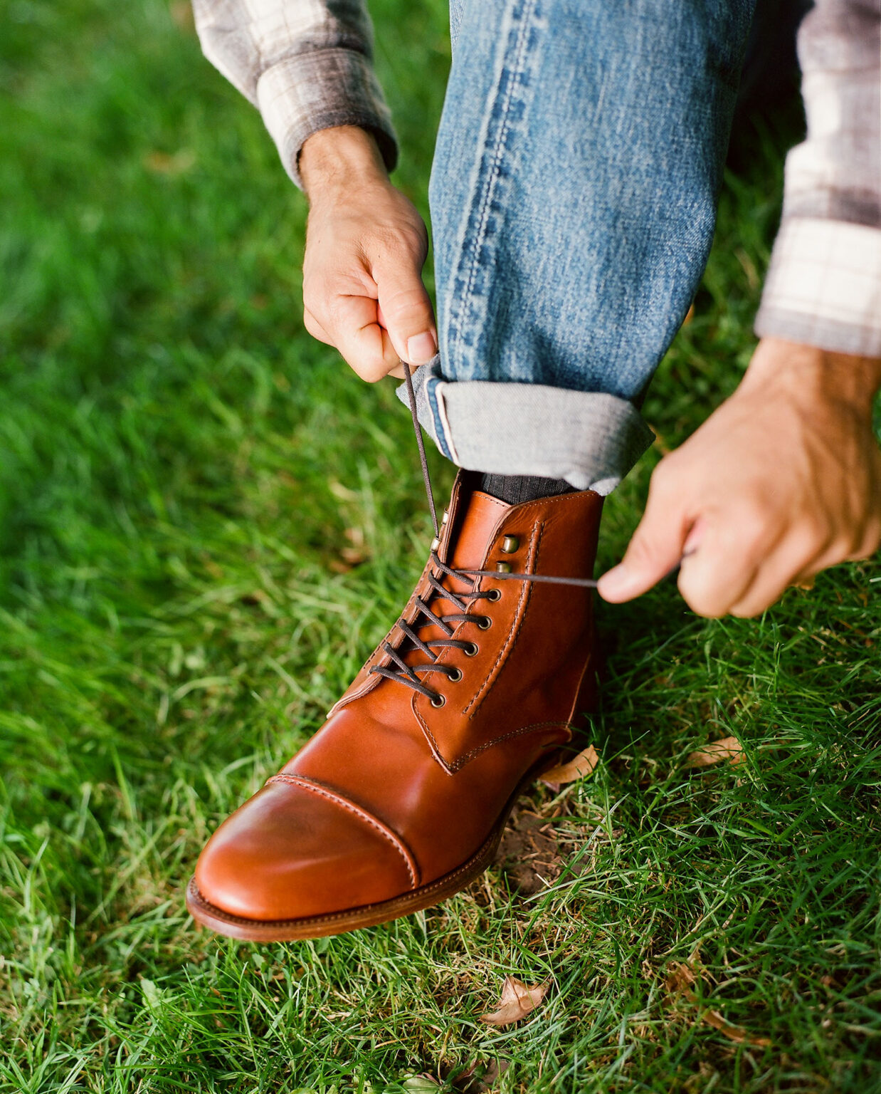 A closeup of a gentleman in a field wearing turned up denim jeans tying up the laces of an E.Woodford Barrack 'Stanley' WWII Boot in Chestnut Spanish Veg Tanned Aniline Calf