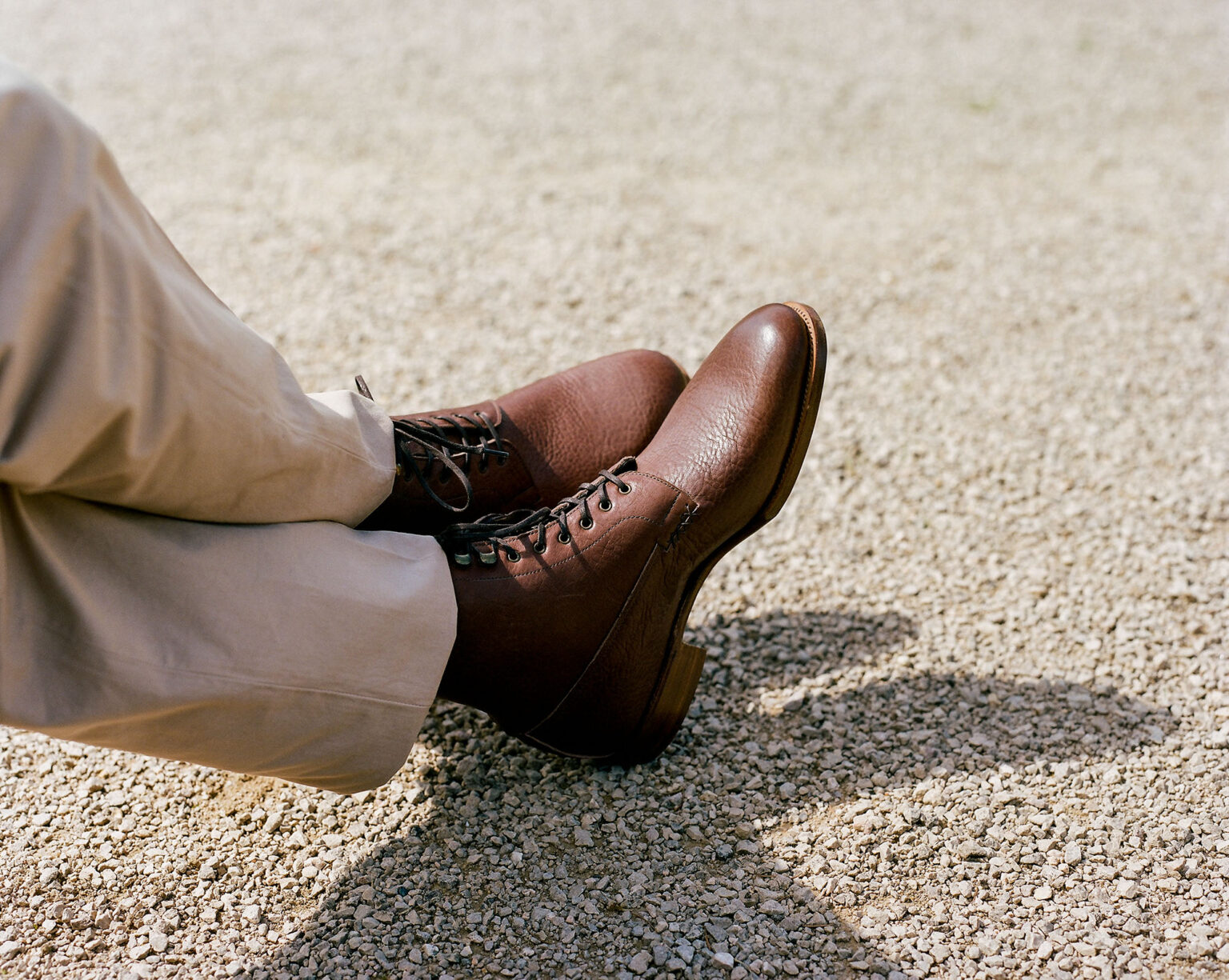 Close up of a gentleman's legs resting on gravel wearing beige chinos and E.Woodford Daneville 'Komrade' WWI Boots in Brown Horween Shell Cordovan leather