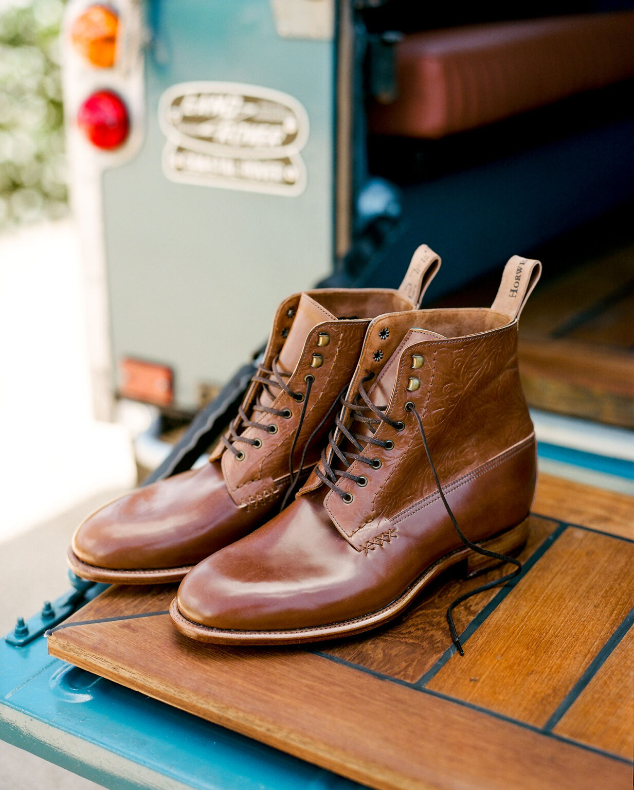 A pair of E.Woodford Daneville 'Komrade' WWI Boots in Bourbon Floral Horween Shell Cordovan resting on the wooden tailgate of a vintage Land Rover Defender in the background