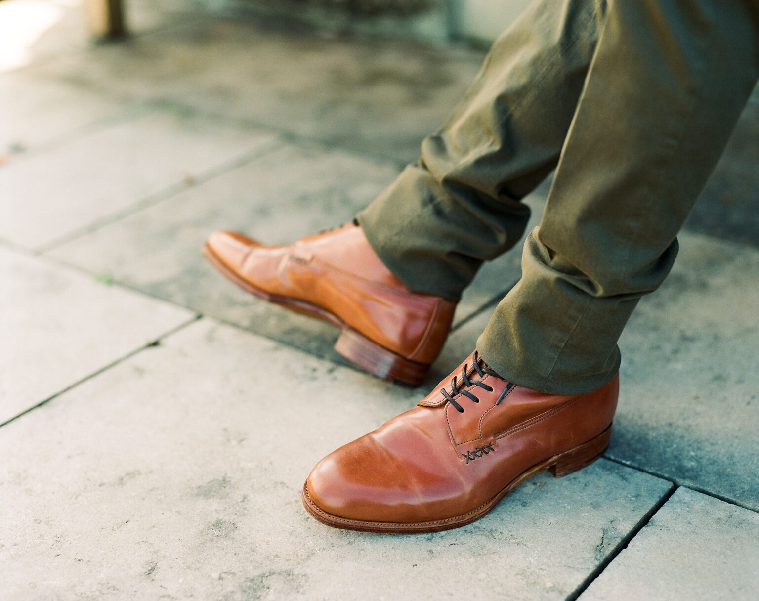 Close up of a gentleman's legs resting on weathered patio slabs wearing khaki chinos and E.Woodford Daneville 'Komrade' WWI Boots in Bourbon Horween Shell Cordovan leather