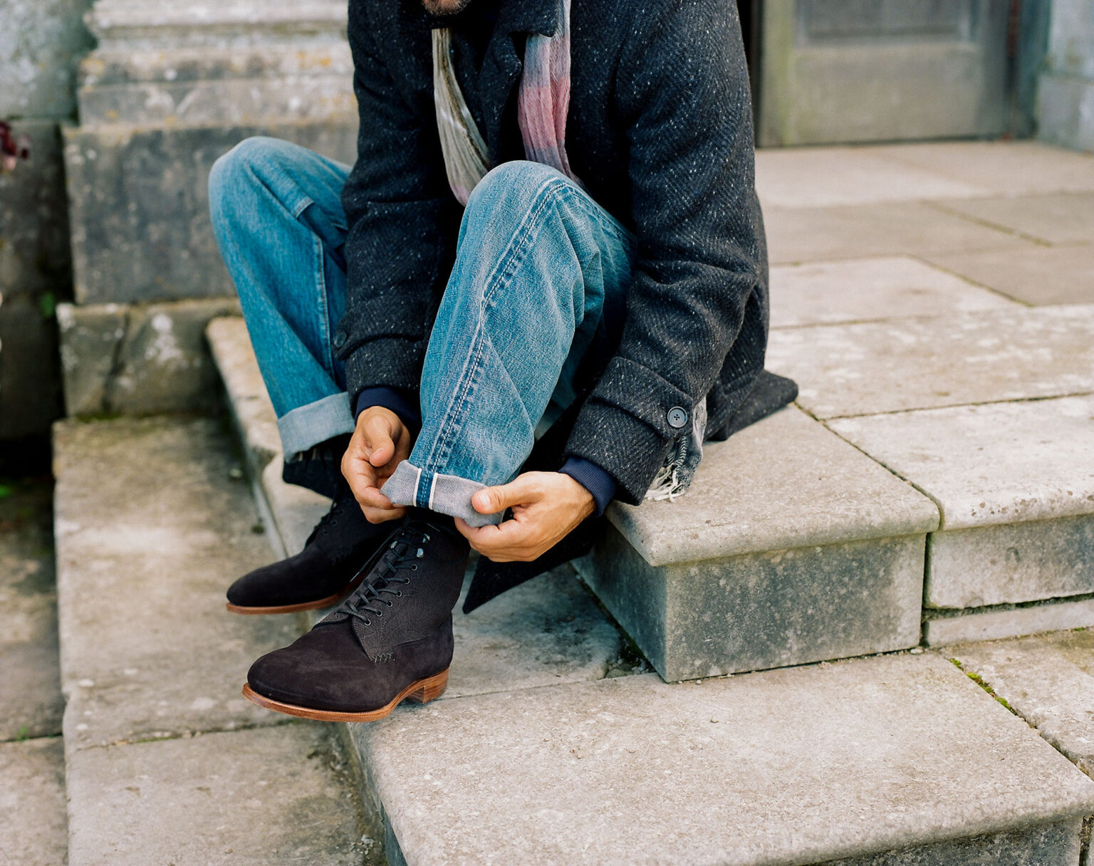 Well dressed man sitting on patio steps turning up the bottom hem on his jeans while wearing a pair of E.Woodford Daneville 'Komrade' WWI Boots in Black CF Stead Janus Calf