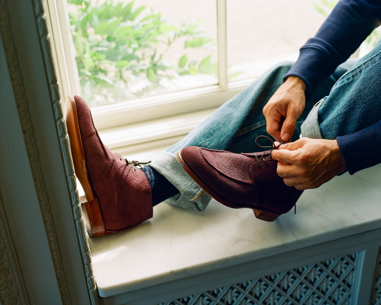 A gentleman sitting on a bay window seat tying laces on a pair of E.Woodford Edgware Ankle Boots in Dark Tobacco CF Stead Janus Calf leather with sunlight flooding in through the window