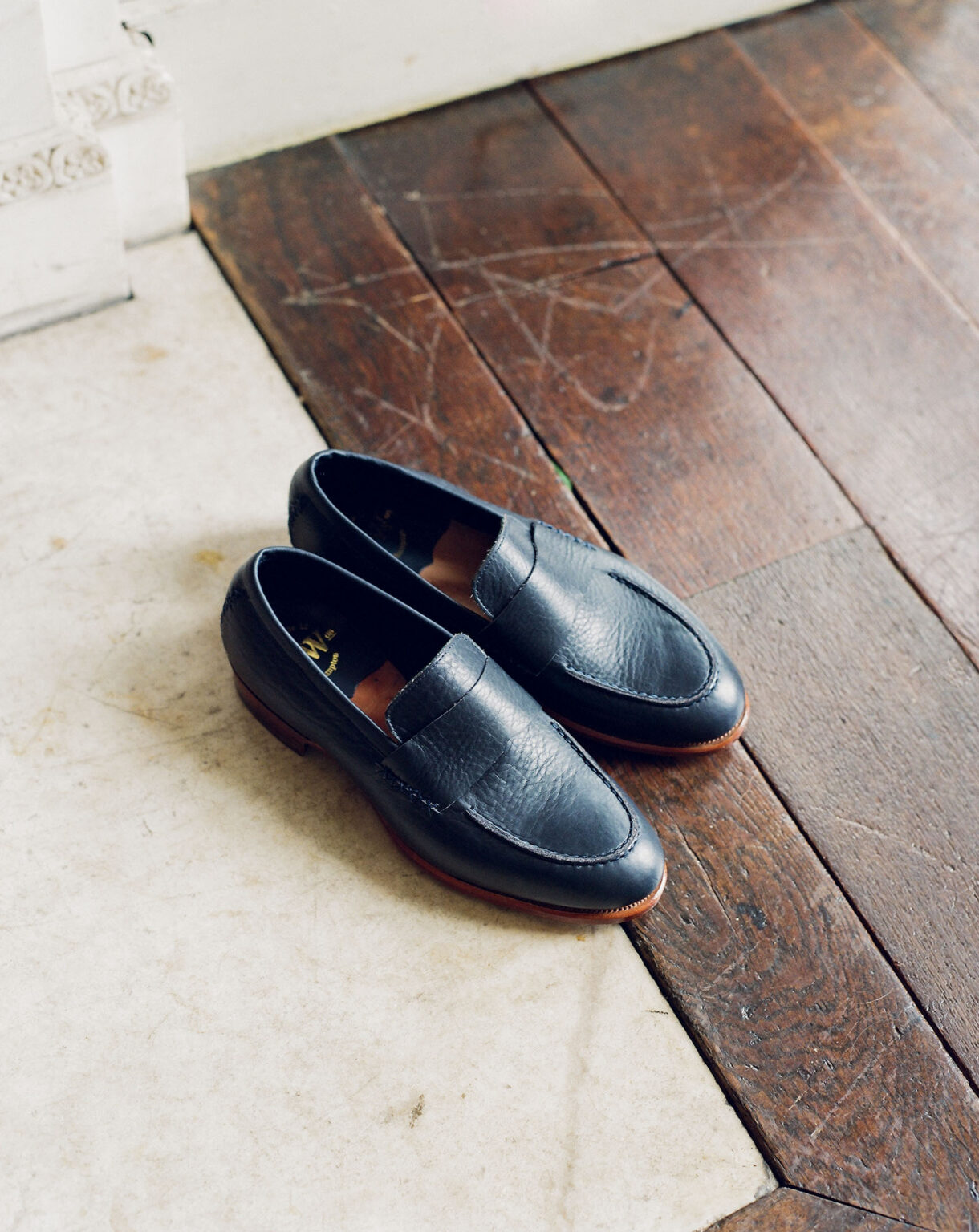 A pair of E.Woodford Fen Baseball Loafers in Navy Horween Veg Tanned Bison laying on a mable and distressed wood floor