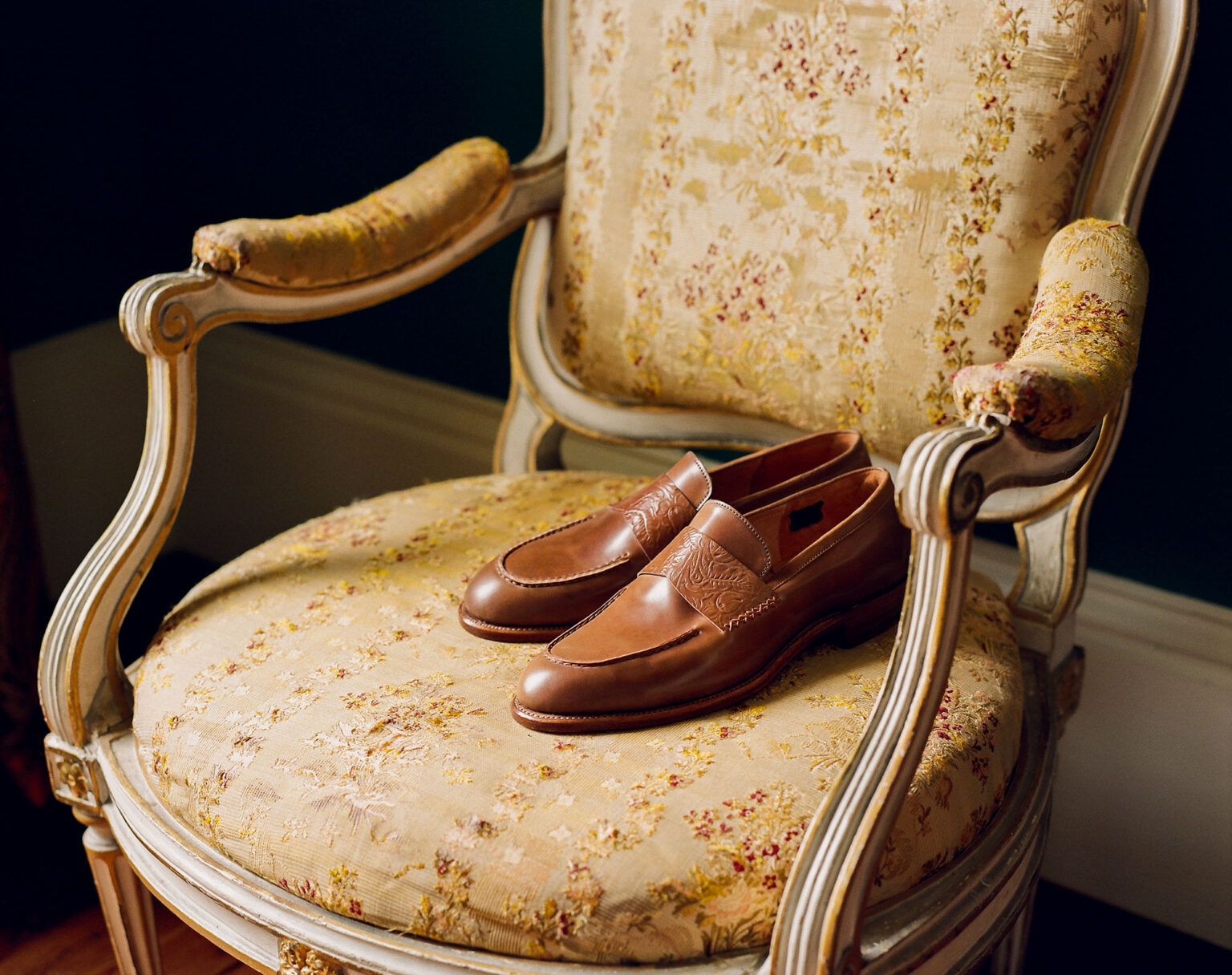 A pair of E.Woodford Fen Baseball Loafers in Bourbon Floral Horween Shell Cordovan sitting on a French Palais Royal armchair