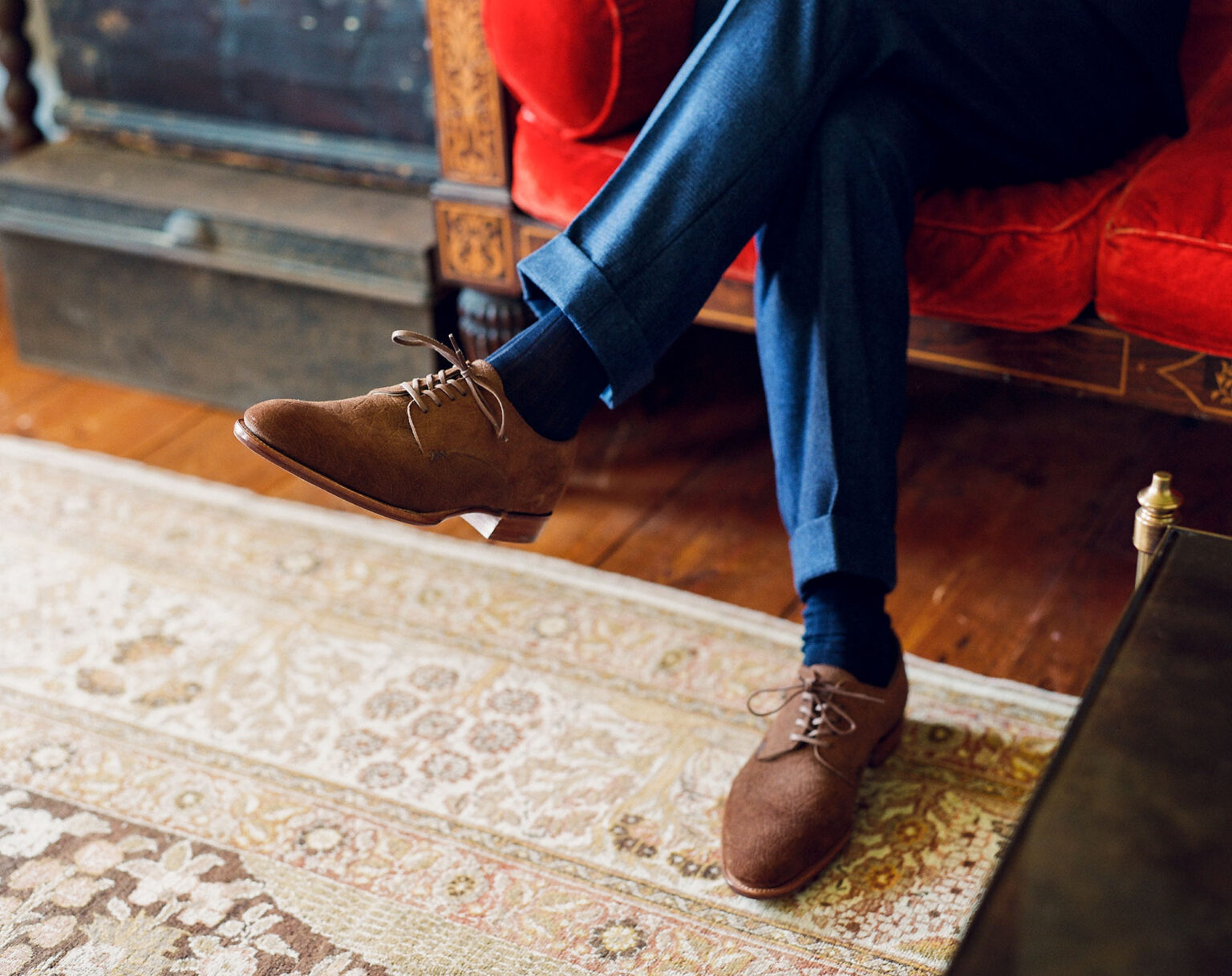 Closeup of gentleman sitting on an ornate red sofa wearing a pair of turned up navy blue tweed suit trousers and a pair of E.Woodford Kennington Gibsons in Dark Chestnut CF Stead Janus Calf with Ernest emboss