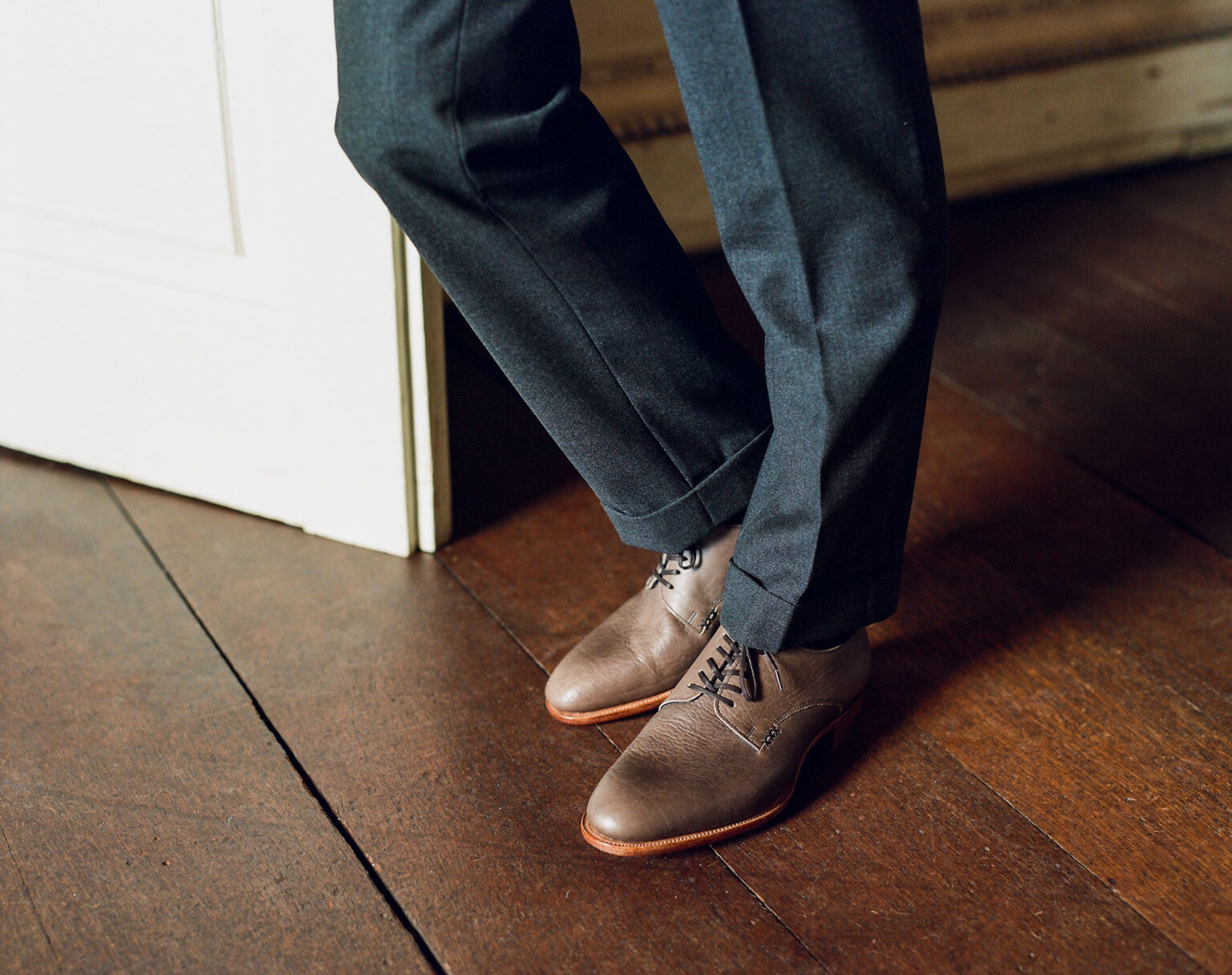 Closeup of lower half of gentleman's legs standing in a doorway on an old oak floor wearing grey suit trousers and a pair of E.Woodford Kennington Gibsons in Grey Horween Veg Tanned Bison