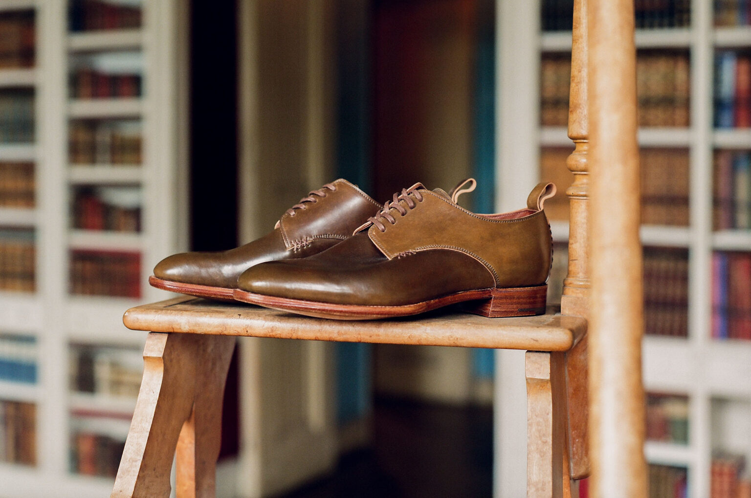 A pair of E.Woodford Kennington Gibsons in Armagnac Horween Shell Cordovan standing on a wooden table in front of home library