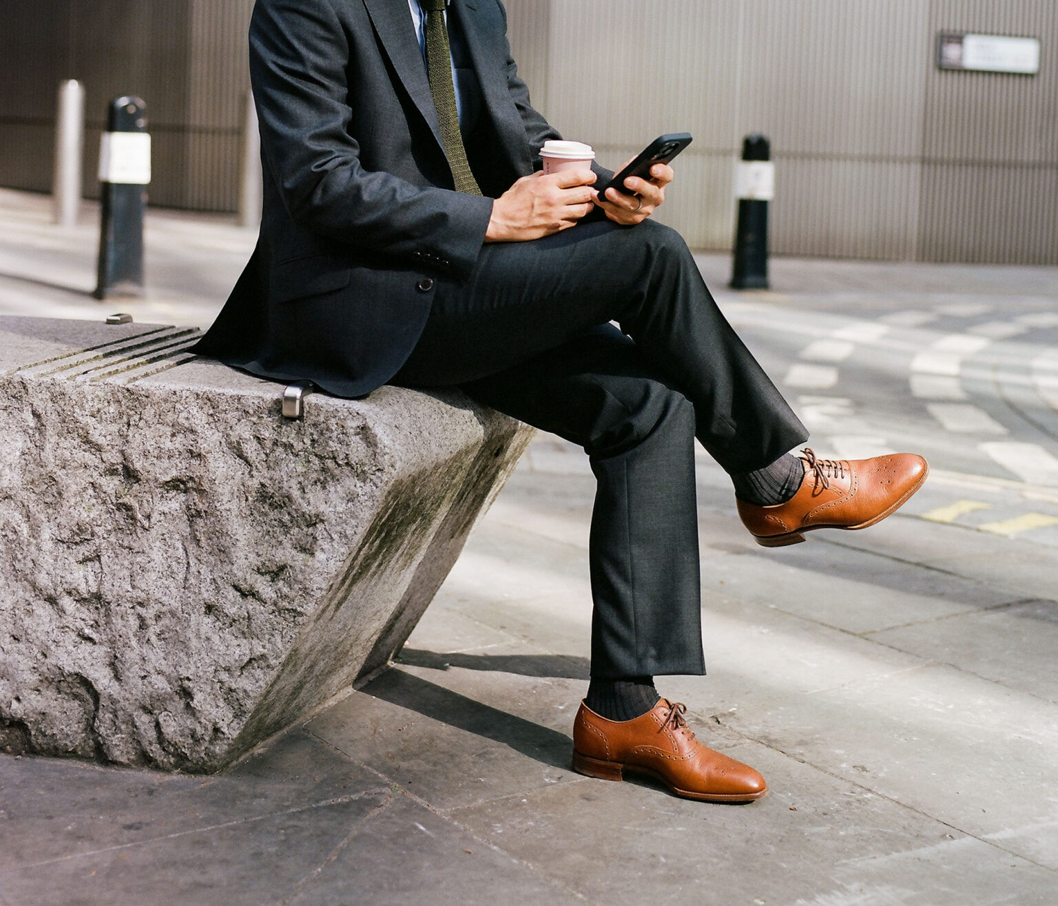 Lower half crop of city gent in a tailored suit sitting down on lunch break holding a coffee and a mobile phone wearing a pair of E.Woodford Redbourne Five Eyelet Oxford shoes in Tan Horween Veg Tanned Bison leather