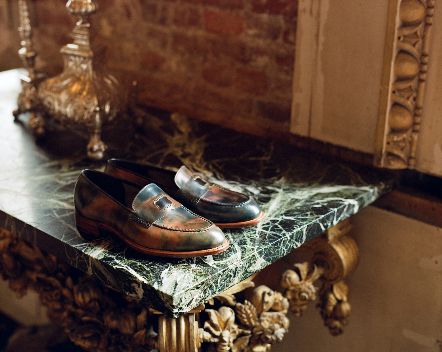 A close up of a pair of E.Woodford Watling Penny Loafers in Black Marbled Horween Shell Cordovan sitting on an ornate marbled surface with gold leaf ornaments in the background