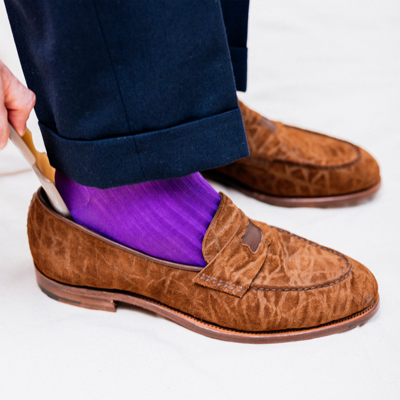 Studio close up of a person using a shoehorn to put on a pair of E.Woodford Watling Penny Loafers in Dark Chestnut CF Stead Janus Calf wearing navy trousers and bright indigo socks