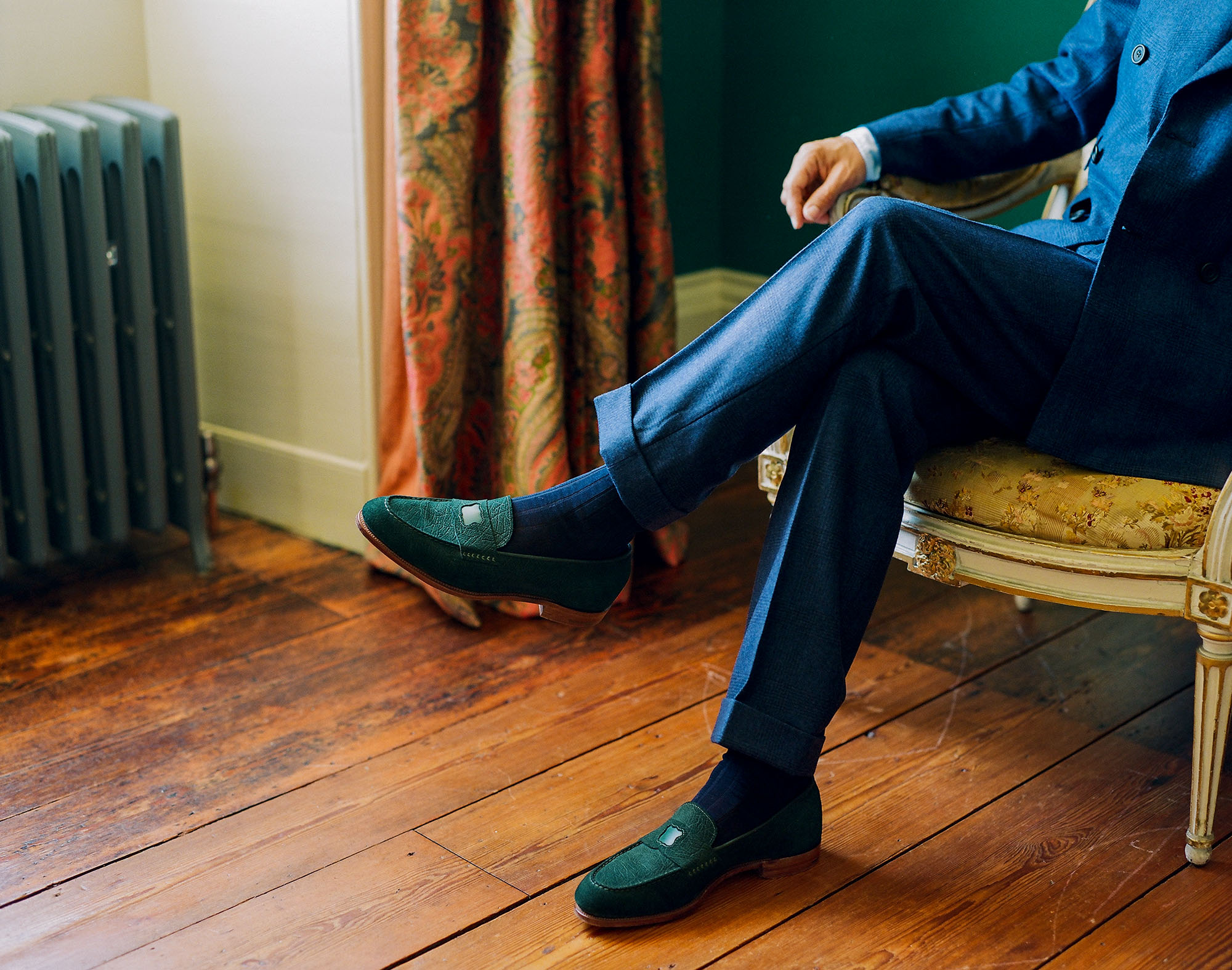 City gent sitting in a french style royal armchair inside a stately home wearing a tailored blue suit with close up focus on E.Woodford Watling Penny Loafers in Racing Green CF Stead Janus Calf