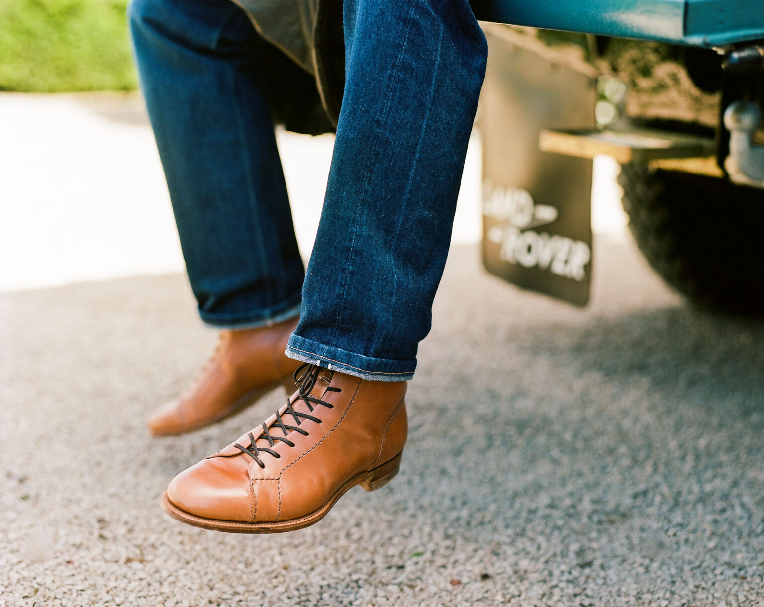 Close up of the legs of a gentleman sitting on the tailgate of a classic Land Rover Defender weating turned up denim jeans and a pair of E.Woodford Woburn Monkey Boots in Armaganc Horween Shell Cordovan leather