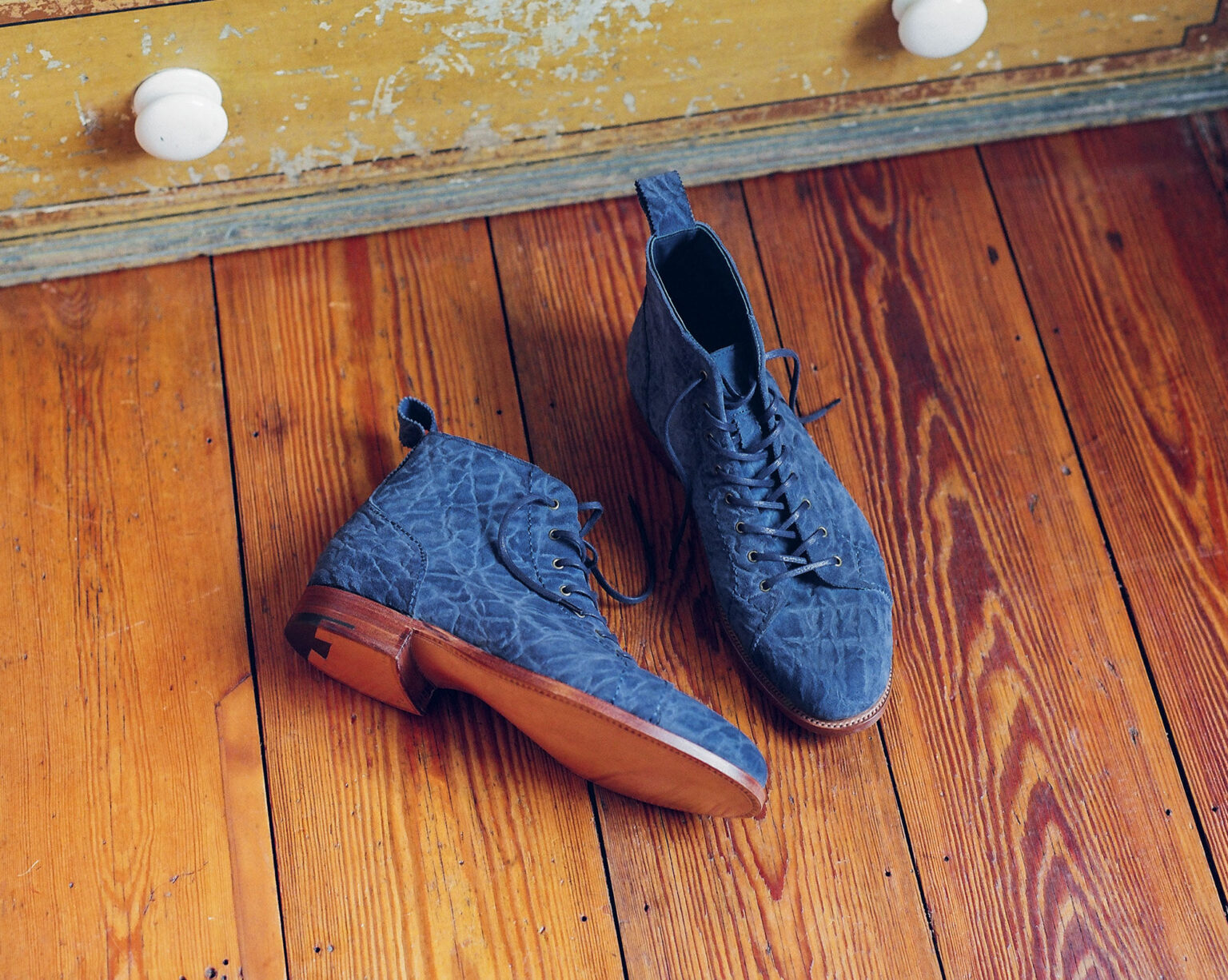 A pair of E.Woodford Woburn Monkey Boots in Navy CF Stead Janus Calf with Edwin emboss on distressed oak floor boards next to a set of shabby drawers with white handles